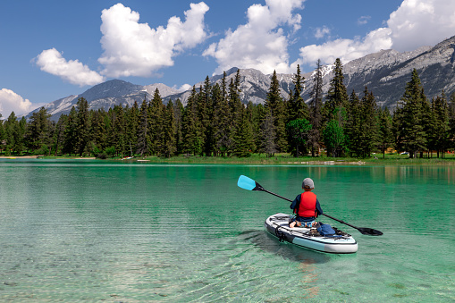 A cute redhead boy is paddleboarding on Edith Lake in Jasper National Park, Alberta. The turquoise colored lake is very calm. The child is sitting on the paddle board in kayak mode. Its is a beautiful sunny summer day in the Canadian Rockies.