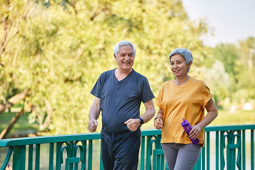 Older Asian and Caucasian wife and husband wear activewear strolling along bridge in summer park, enjoy morning sportive walk together outside. Healthy lifestyle and retirement