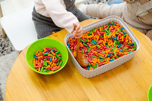 Toddler playing sensory game with colored pasta using bowls and wooden spoons
