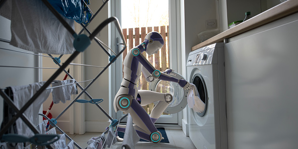 A generic blue and white domestic robot kneeling down and pulling clothing from a washing machine in a domestic utility room with washing hanging up on a drying rack in the foreground. With shallow depth of field.