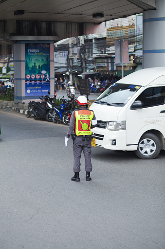 Thai traffic cop is standing at junction on street in Bangkok downtown and is regulating traffic. In background a van is passing. Scene is at junction Ratchadamri Rd and Phetchaburi Rd