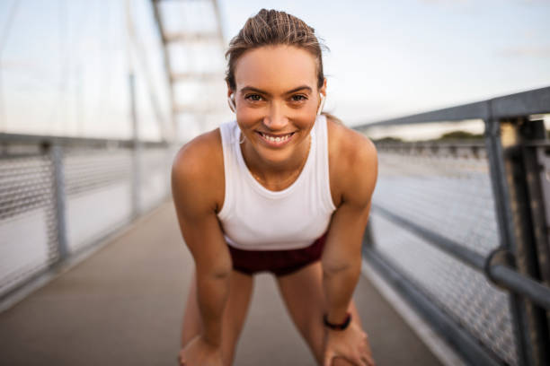 young sportswoman taking a break after running on the bridge looking at camera smiling - sportsman looking at camera full length sport imagens e fotografias de stock