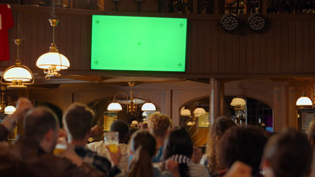 Group of Multicultural Friends Watching a Live Sports Match on TV with Green Screen Display in a Bar. Happy Fans Cheering and Shouting, Celebrating When Team Scores a Goal and Wins the Tournament.