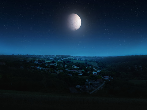 Huge moon in the starry sky above the hills. Moonlight illuminates the night landscape with a town on a hill.