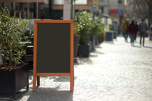 An empty menu board near a stylish outdoor cafe in a European city on the background of urban street. Off-season. Outdoor restaurant. Mockup for designer.