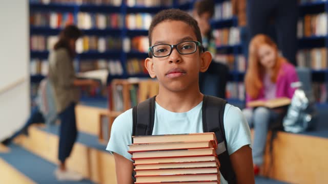 Middle eastern boy holding stack of books against multi colored bookshelf in library.