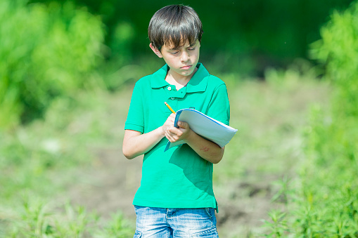 A young elementary student walks with his science class outdoors as they learn about the plants and creatures around them.  He is dressed casually as he walks with a piece of paper and a pencil to take notes as he observes his surroundings.