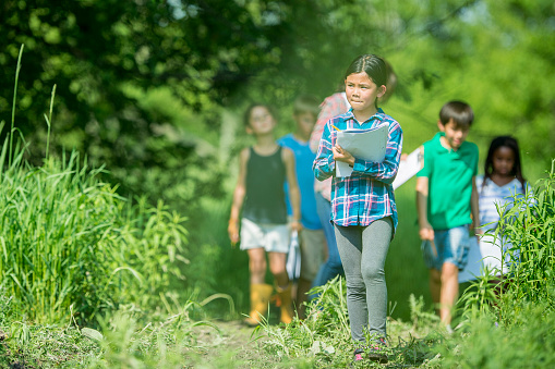 A small group of elementary students take their science class outdoors as they learn about the plants and creatures around them.  They are each dressed casually as they walk with clipboards and take notes.