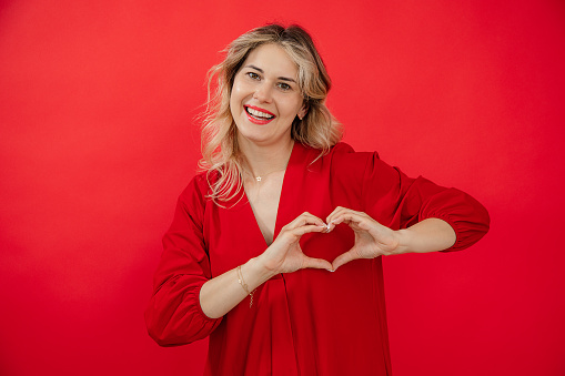 Portrait of joyful attractive young woman in holiday red look with perfect bright makeup isolated on red background. Love expression, making heart shape by hands. Fashion and beauty industry.