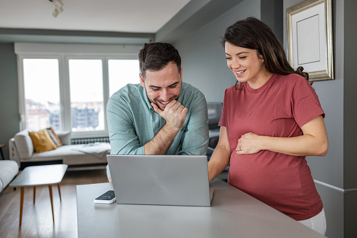 Lovely pregnant couple watching videos about pregnancy on the internet. They are smiling while looking at the laptop. Preparation for parenting.