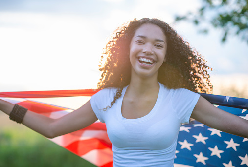 A young female teenager of African decent holds a USA flag in her hands as it fans in the air behind her.  She is dressed casually in a white -t-shirt and is smiling as she shows her patriotism.