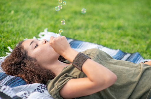 A young teenager of African decent lays on a picnic blanket in the warm sun as she holds a Bubble wand and blows gently.  She is dressed casually and is enjoying her leisurely time alone.