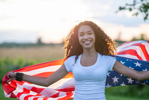 A young female teenager of African decent holds a USA flag in her hands as it fans in the air behind her.  She is dressed casually in a white -t-shirt and is smiling as she shows her patriotism.