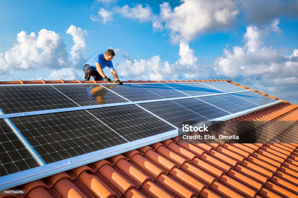 Professional worker installing solar panels on the roof of a house kneeling professional fixing solar panels from the top of a house roof, side view of the roof with sun reflection Solar Panel Stock Photo