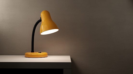 Stylish glowing yellow desk lamp on white table in room, copy space for text on gray plaster wall background.