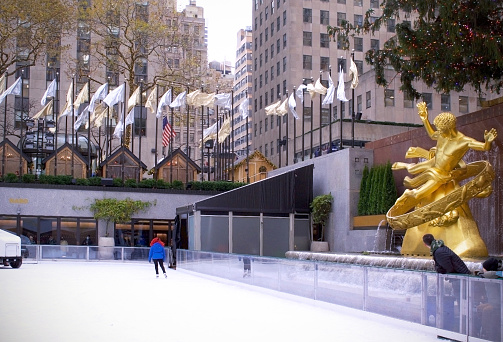 New York, NY, USA-December 6, 2022: Holiday tradition and recreation-Rockefeller Center renovated buildings allow views around the rink at the rink level of this complex. Seen here- a skater with a boy, a man, and a statue of Promethius looking on