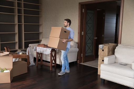 Young single man carrying big packed cardboard boxes with personal belongings inside unfurnished living room on relocation to new first own or rented house. Homeowner move day, tenancy, bank mortgage