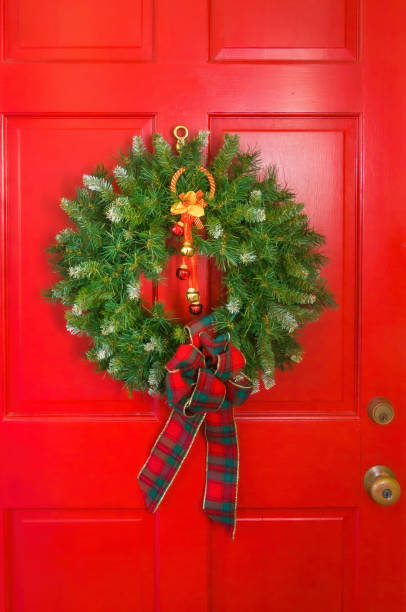 A green Christmas wreath with a tartan plaid ribbon hangs on a red door of a Cape Cod home.