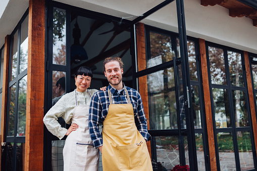 Cheerful smiling man and woman standing at the entrance door. They are wearing aprons for work as barista and a waiter in a coffee shop. They are looking at the camera.