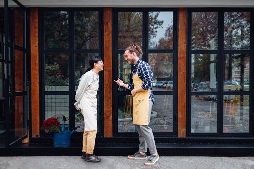 Handsome smiling man with long hair and beautiful Asian woman working in a cafe, standing at the entrance door. They are wearing aprons for work as barista and a waiter. They are looking at each other and smiling.