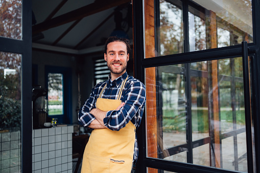 Portrait of a handsome confident man working as a barista and wearing an apron. He is standing at the entrance of his cafe and looking at camera while smiling.