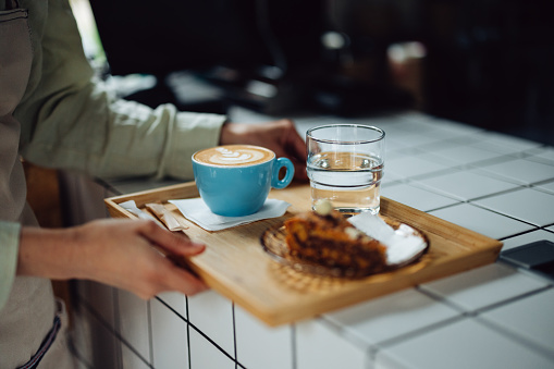Unrecognizable woman carrying a tray with a cup of coffee, plate with a piece of cake and a glass of water. She is working in a cafe.