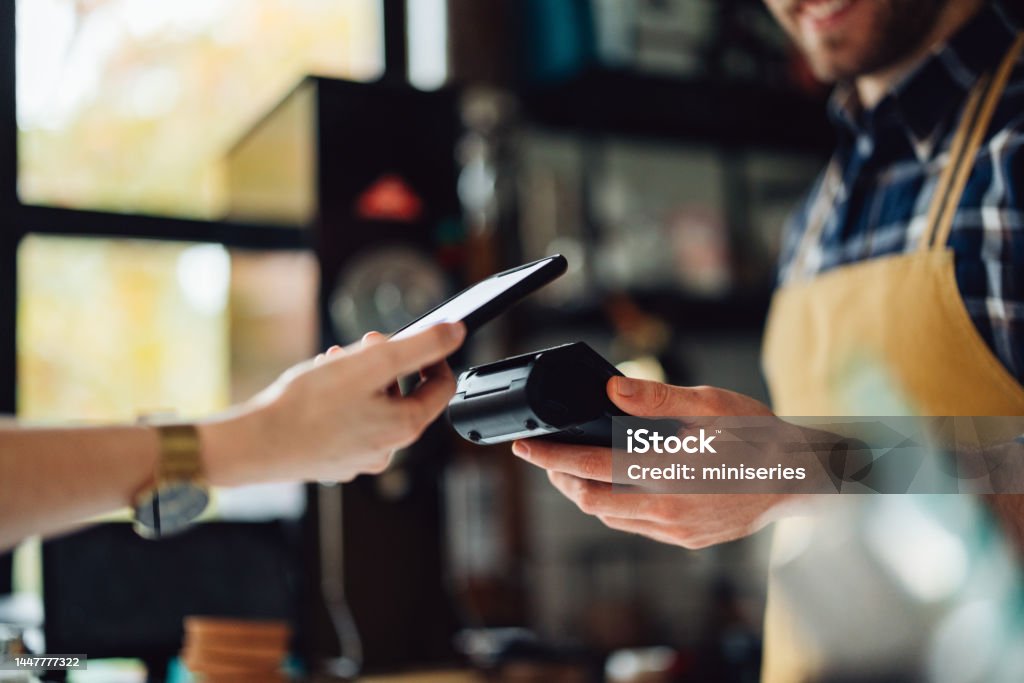 Anonymous Person Paying With Their Cell Phone Unrecognizable people making an online payment. Anonymous man is holding a card machine while the other person is placing their phone to pay. Paying Stock Photo