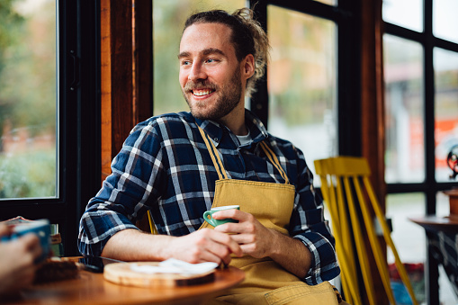 Handsome cheerful man sitting at the table and drinking coffee. He is smiling while looking away.