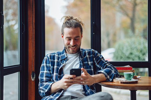 Portrait of a cheerful man with long hair in a bun, sitting in a cafe by the window. He is smiling while looking down and texting on his mobile phone.