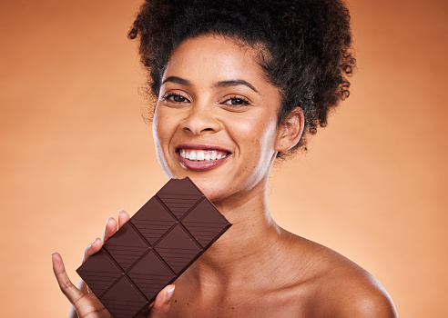 Chocolate, happy woman and beauty portrait on studio background eating sweets, dessert or sugar. Young african american girl bite dark chocolate bar and cocoa candy, calories and delicious food snack