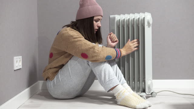 A woman freezes at home near the heater, suffers from frequent power outages. Shutdowns of electricity and central heating due to the energy crisis.