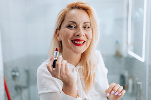 Adorable mid adult Caucasian blonde woman with a gorgeous big smile applying lipstick in bathroom