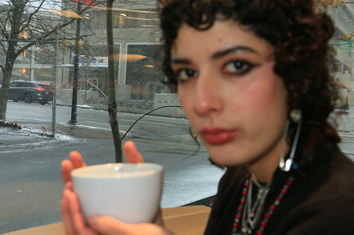 An Hispanic woman drinking hot tea while watching a snowstorm in Autumn outside. She is wearing a black shirt, necklaces and earrings.