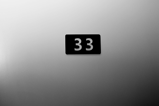 Hotel room 33 number mounted on a white wall. White number on black board by wooden doors in corridor hall.