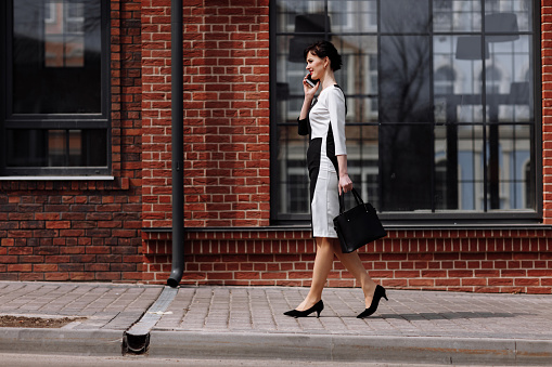 Happy lawyer businesswoman professional is walking outdoors talking on cell smart phone. Caucasian successful female is smiling wearing stylish white-black dress with black bag.