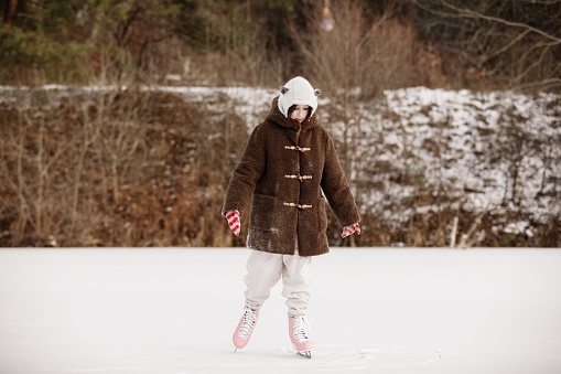 Child ice skating on frozen lake with in the snowy forest. Little girl wearing in warm clothes and pink skates at the rink on natural ice on cold winter day. Happy holiday vacation.