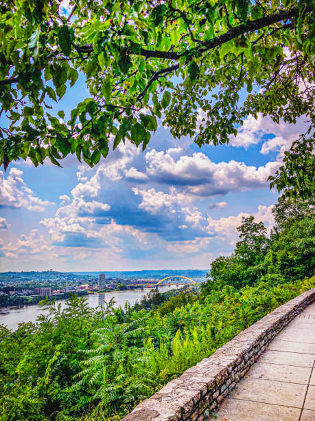 A sidewalk is surrounded by lush foliage in Eden Park Cincinatti Ohio.  The scene overlooks Covington Kentucky on a partly cloudy summer day. There are downtown buildings in the distance and a bridge spans across the mighty Ohio River.