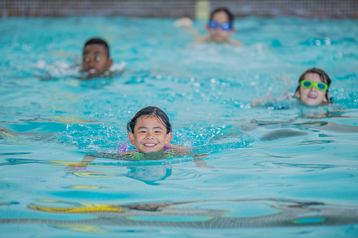 A small group of school-aged children are seen swimming in an indoor pool during a lesson.  They are each wearing swimsuits and smiling as they tread water.