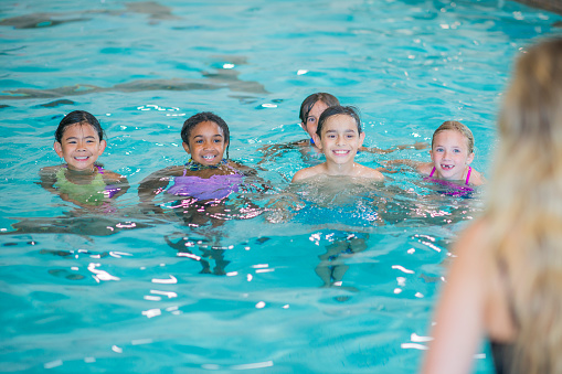 A small group of school-aged children are seen swimming in an indoor pool during a lesson.  They are each wearing swimsuits and smiling as they tread water.