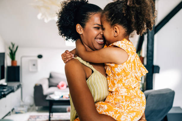 A Beautiful Cute Girl Hugging Her Smiling Mother While She Is Holding Her A side view of a pretty African-American child embracing her loving nanny while they are having fun at home. excited woman yellow stock pictures, royalty-free photos & images