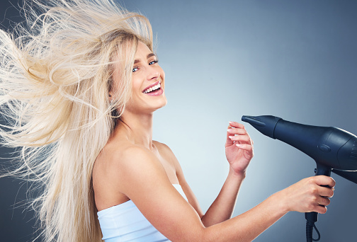Beauty, hair care and woman on blue background with hair dryer, smile and healthy blonde salon hair style. Health, wellness and luxury care for happy model hair, color treatment, shampoo and dry hair
