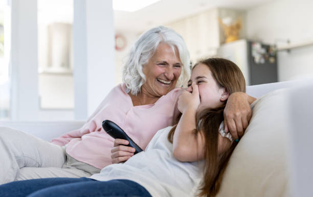 Happy teenage girl laughing while watching tv with her grandmother at home Happy teenage girl laughing while watching tv with her grandmother at home - family lifestyle concepts teenage girl with grandmother stock pictures, royalty-free photos & images