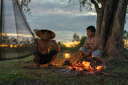 Asian village lifestyle, a local male fisherman wearing conical hat with his son drinking a cup of coffee fixing his fishing net under a tree beside a campfire and freshwater lake during dawn sunrise time, very beautiful scenic view