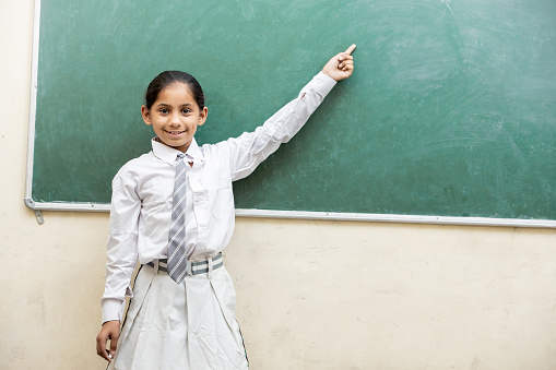 Happy indian school girl kid pointing on blank chalkboard or blackboard while standing in classroom. female Education concept.