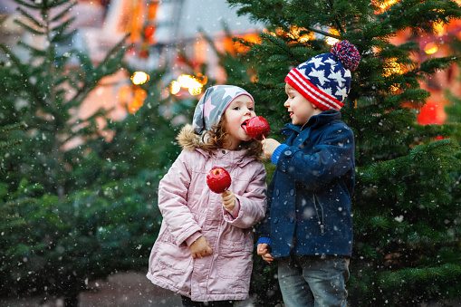 Two little smiling kids, boy and girl eating crystalized sugared apple on German Christmas market. Happy friends in winter clothes with lights on background. Family, tradition, holiday concept.