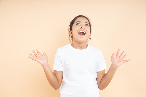 Amazed little indian girl looking up with open arms gesture isolated over beige background. Imagination and day dreaming concept.