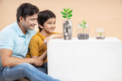 Growing Money and Investment Concept - Happy Young Indian Father and Song Sitting With Coin In Glass Jar With Plant Grow In It.