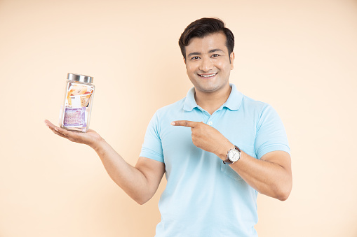 Portrait of happy Indian man pointing at glass jar full of rupee notes isolated on beige studio background. Money saving and investing concept.
