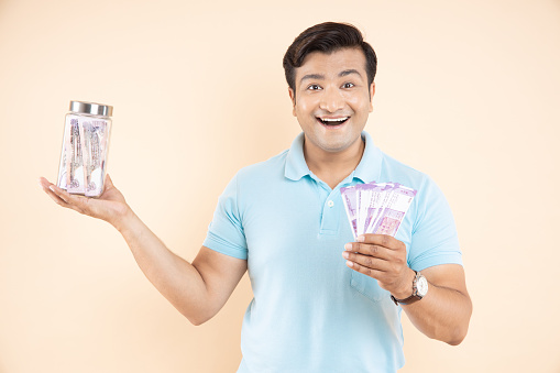 Amazed or cheerful handsome man with Indian rupee notes in one hand and glass jar with lots of cash money in other hand isolated on beige studio background. saving and smart investing concept.
