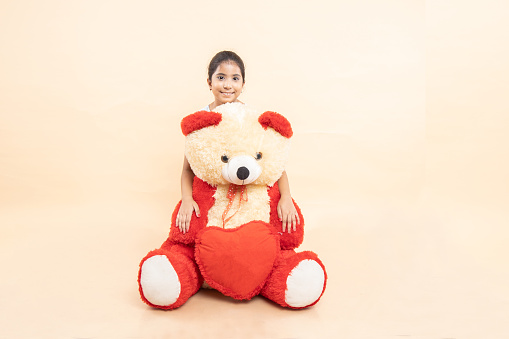 Happy indian girl kid hugging her big teddy bear isolated over beige studio background.